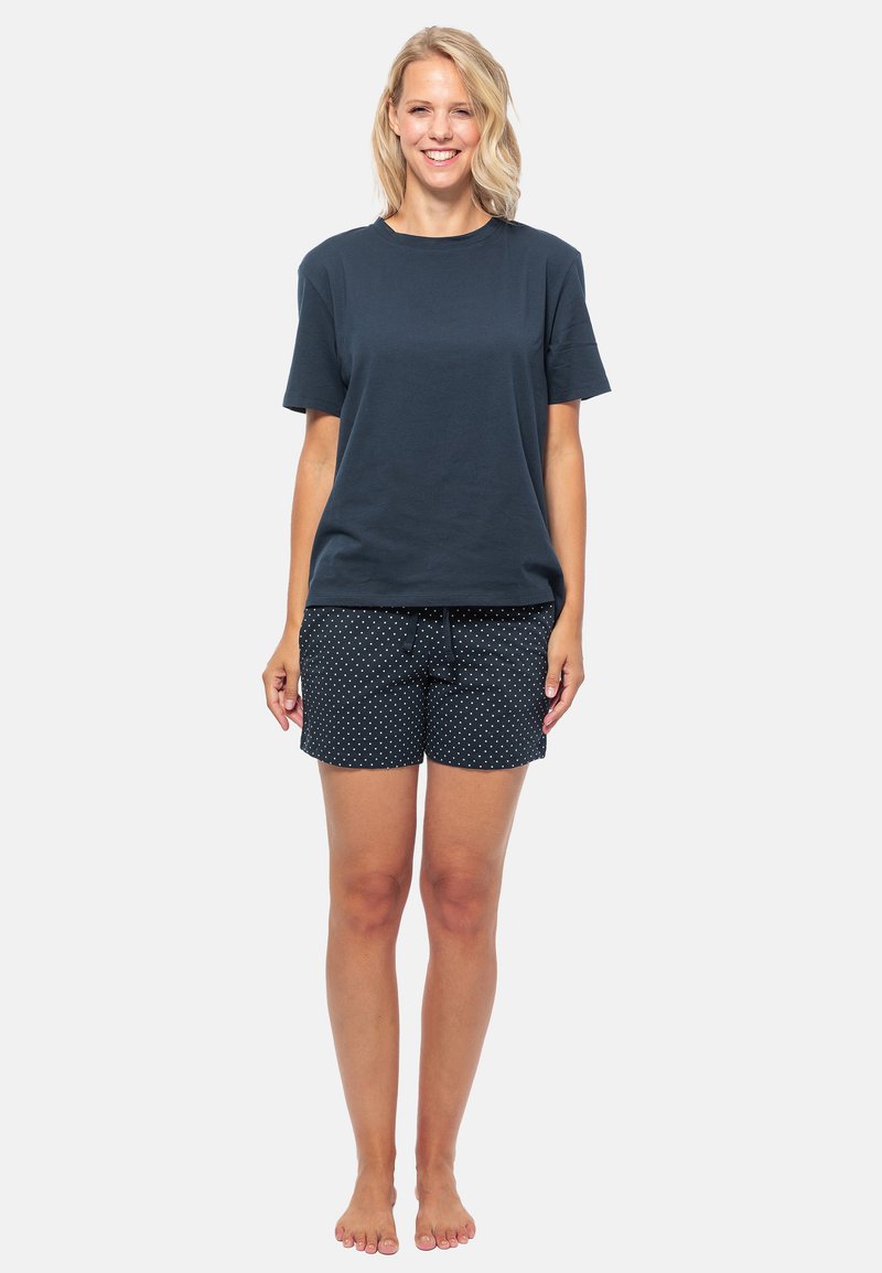 Marineblauwe katoenen t-shirt met korte mouwen, gecombineerd met zwarte shorts met witte stippen. Model staat op blote voeten tegen een effen achtergrond.