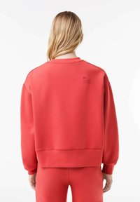Röd sweatshirt med rundad hals, ribbade ärmslut och nederkant. Lacoste-logotyp tryckt på den övre delen av ryggen. Slät tygtextur.