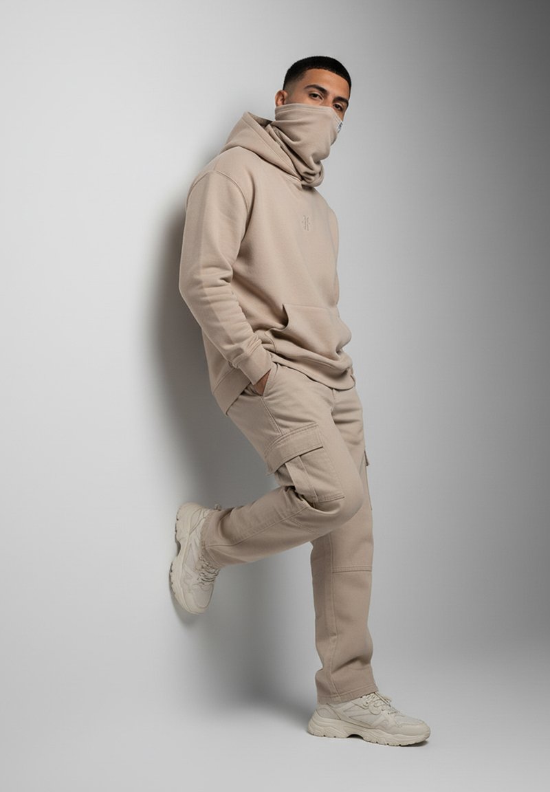 Beige hoodie with high neck and kangaroo pocket, paired with beige cargo pants featuring side pockets. White sneakers complete the outfit.