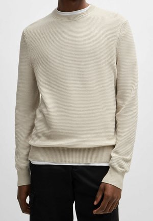 Mężczyzna ubrany w kremowy, prążkowany sweter z dekoltem typu crewneck na białej podkoszulce, w czarnych spodniach, stojący na jednolitym tle.