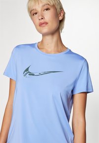 Camiseta deportiva azul claro hecha de una tela suave, con un logo de Nike verde oscuro en el frente. Manga corta y escote redondo.