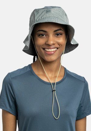 Persona sonriente que lleva un sombrero tipo cubo Buff de color gris claro con correa ajustable para la barbilla y una camiseta deportiva azul de manga corta sobre fondo blanco.