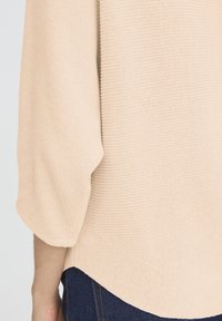 Beżowy sweter z dzianiny o prążkowanej fakturze, z rękawami do łokcia i zaokrąglonym dołem. Połączony z ciemnoniebieskimi dżinsami.