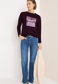 Bordeauxfarbendes Langarmshirt mit bedrucktem Text, kombiniert mit blauen Schlagjeans. Das Model hält eine cremefarbene, strukturierte Jacke.