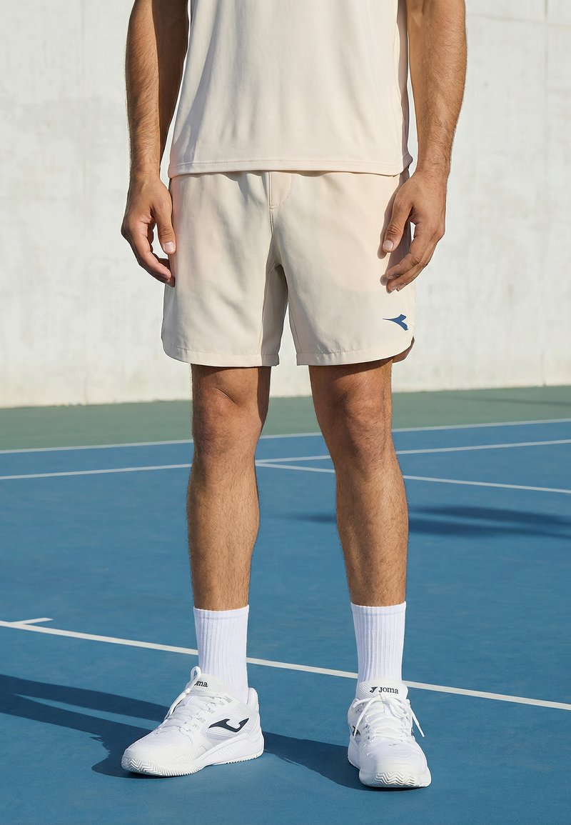 Mand står på udendørs tennisbane iført beige shorts, hvide crew-strømper og hvide sportssko med logo.