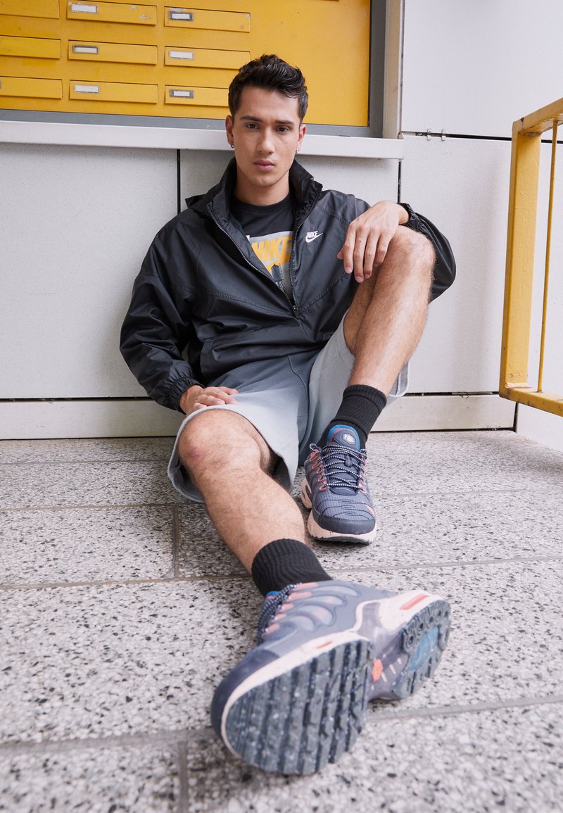 Nike melns vējjakas, pelēkas šortus un tumšas sporta kedas ar rozā akcentiem. Sēž uz teksturēta grīdas netālu no dzeltenajiem glabāšanas skapjiem.