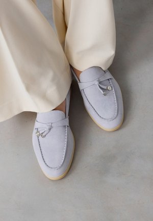 Mocassins en daim gris clair avec pompons décoratifs portés avec un pantalon beige à jambes larges sur un sol gris lisse.