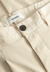 Pantalon beige en mélange de coton avec une texture côtelée, doté d'un bouton foncé et d'une étiquette "GABBA" sur la ceinture. Détails de couture visibles.