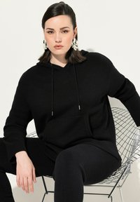 Felpa nera oversize realizzata in tessuto a maglia con tasca frontale, cappuccio regolabile con cordino e polsini a costine, abbinata a leggings neri coordinati.