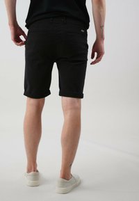Shorts en denim noir avec des revers roulés, des poches arrière et une étiquette. Portés avec des chaussures à enfiler de couleur claire. Fond neutre.