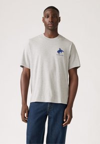 Grijze katoenen t-shirt met korte mouwen, ronde hals en blauw logo op de borst. Gecombineerd met donkerblauwe spijkerbroek.