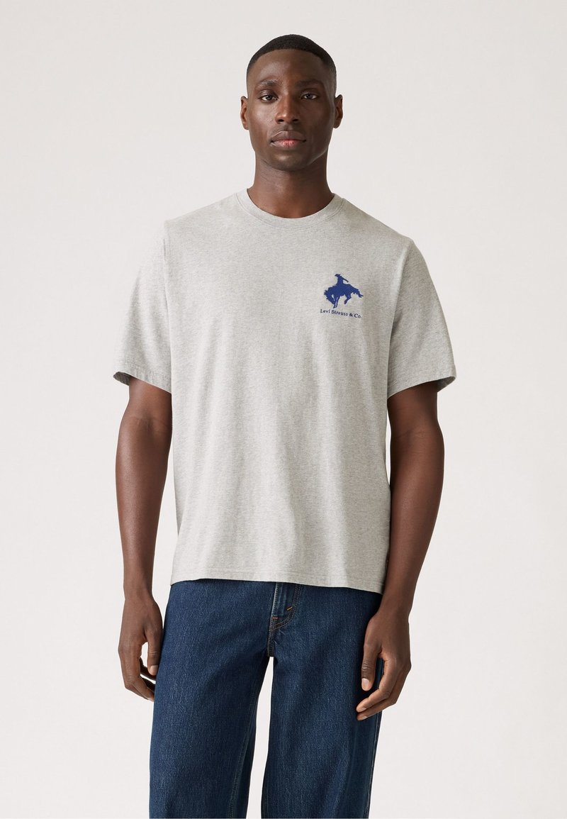 Grijze katoenen t-shirt met korte mouwen, ronde hals en blauw logo op de borst. Gecombineerd met donkerblauwe spijkerbroek.