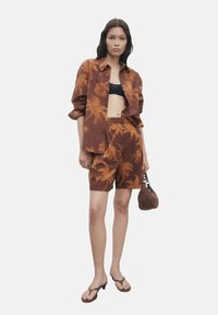 Set di camicia marrone e pantaloni corti con motivi di palme, realizzato in tessuto leggero. Un crop top nero e una borsa marrone completano l'outfit.