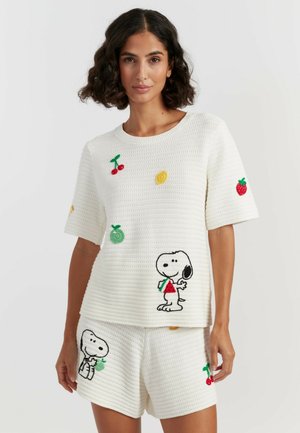 Donna che indossa pantaloncini e top bianchi in maglia con patch di Snoopy e frutta, tra cui ciliegie, limone, fragola e mela.