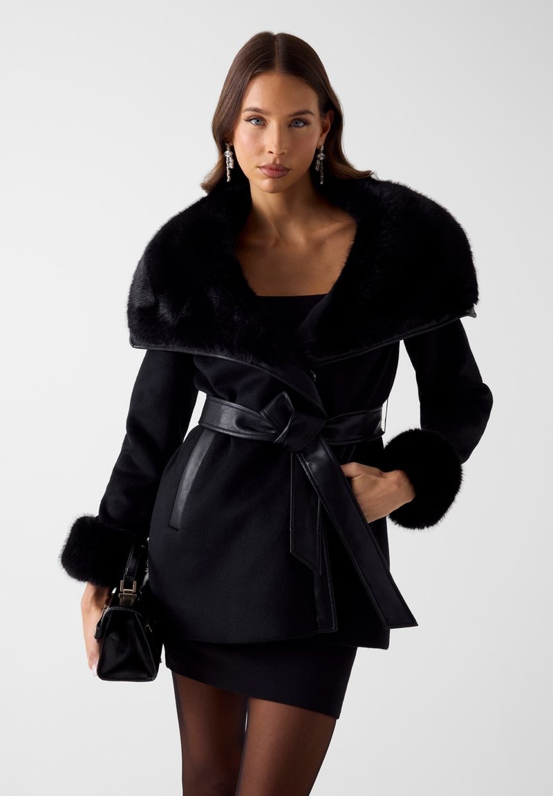 Zwarte jas met brede bontkraag, leren riem en bontmanchetten. Gestructureerd ontwerp, gladde textuur, gecombineerd met een zwarte mini-jurk en handtas.