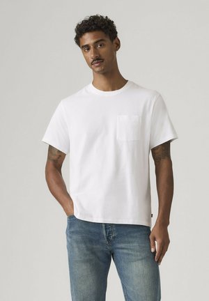 Mann mit kurzen lockigen Haaren und Schnurrbart, der ein weißes T-Shirt mit kurzen Ärmeln und Brusttasche sowie blaue Jeans trägt, steht vor einem einfarbigen Hintergrund.