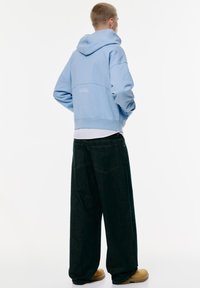Haut à capuche bleu clair avec une coupe décontractée, des poignets côtelés et un détail de logo au dos, associé à un pantalon en denim foncé ample et des bottes beiges.