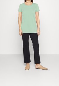 Vaaleanvihreä lyhythihainen t-paita yhdistetty mustiin suoriin housuihin ja vaaleisiin kudottuihin slip-on-kengiin, taustalla yksivärinen tausta.