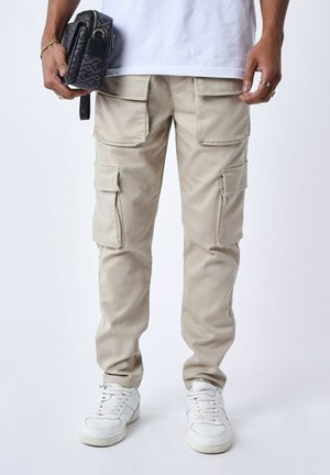 Pantaloni cargo beige con più tasche, vestibilità slim, realizzati in tessuto leggero, abbinati a sneakers bianche e con una borsa nera a motivo.