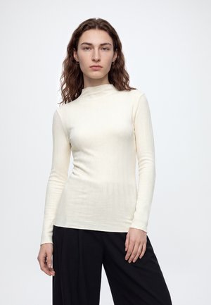 Jeune femme aux cheveux bruns ondulés portant un haut à manches longues côtelé crème et un pantalon noir, debout devant un fond blanc uni.