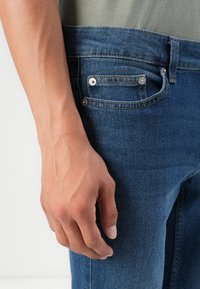 Jeans de denim azul con un diseño ajustado, que cuentan con herrajes plateados y un pequeño bolsillo frontal. Primer plano de una mano descansando sobre el muslo.