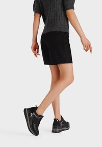 Persona che indossa pantaloni corti neri, un maglione corto grigio scuro e sneakers con la piattaforma nere, che cammina su uno sfondo semplice.