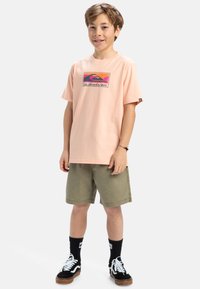 Ljusrosa bomulls-T-shirt med en färgglad grafisk design, kombinerad med olivgröna shorts, svarta strumpor och svarta sneakers med vita detaljer.