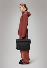 Mala preta de rolo com um bolso à frente, segurada à mão. Pessoa vestindo um casaco impermeável na cor ferrugem e calças combinando.
