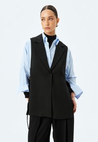Chaleco negro ajustado con un cierre de bot�ón único, usado sobre una camisa azul claro. Presenta solapas anchas y una textura suave. Diseño sencillo.