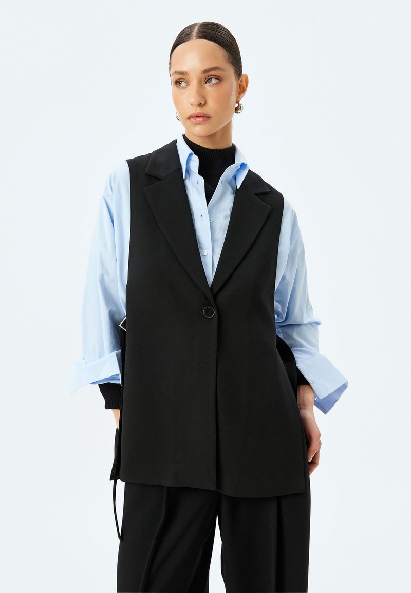 Chaleco negro ajustado con un cierre de botón único, usado sobre una camisa azul claro. Presenta solapas anchas y una textura suave. Diseño sencillo.