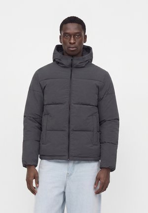 Homme portant une doudoune à capuche gris foncé avec des poches avant et un jean bleu clair, debout devant un fond blanc uni.