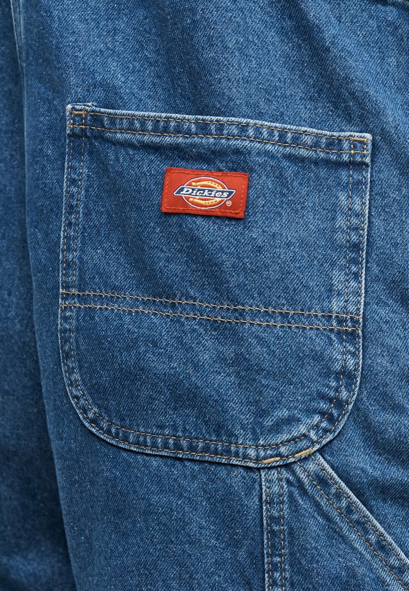Primo piano di una tasca in denim blu con una patch rossa del logo Dickies cucita al centro in alto.