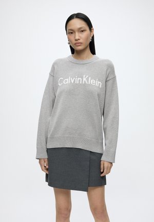 Młoda kobieta z długimi ciemnymi włosami, nosząca perłowe kolczyki, szary sweter Calvin Klein i ciemnoszarą spódnicę na jednolitym jasnym tle.