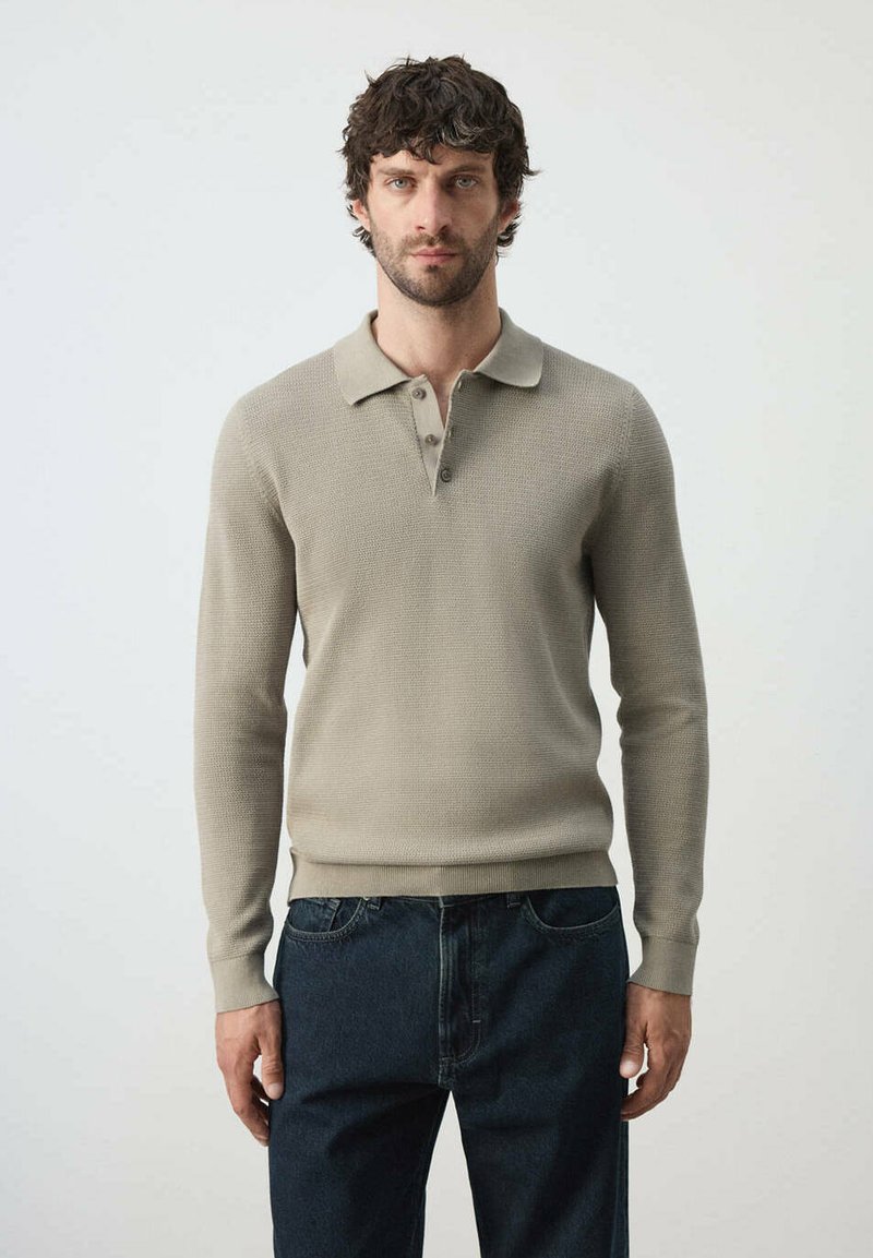 Homme aux cheveux bouclés foncés portant un polo en maille beige à manches longues et un jean bleu foncé, debout devant un fond clair uni.