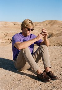 Jeune homme portant des lunettes de soleil, une chemise violette et un pantalon beige, assis en tailleur sur un sol désertique rocheux avec des collines en arrière-plan.