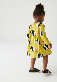 Helles gelbes Kleid mit Panda-Print und floralen Akzenten. Lange Ärmel und taillierter Schnitt. Schwarze Glitzer-Sneaker mit metallischen Details.