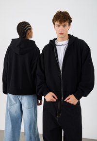 Sudadera negra con cremallera y una capucha grande, de corte holgado y puños acanalados, llevada sobre una camiseta blanca. Combinada con pantalones vaqueros de mezclilla azul claro y oscura.
