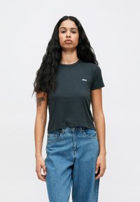 Svart avkortet t-skjorte med rund hals og korte ermer, med en liten hvit "obey"-logo på venstre bryst. Brukt med blå jeans.