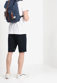 Granatowy plecak z brązowymi skórzanymi akcentami, prostokątny kształt, regulowane paski, noszony z białą koszulą i czarnymi szortami.