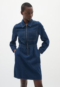 Denim jurk met een voorrits, kraag, lange mouwen, twee borstzakken en een elastische taille voor vormgeving. Donkerblauwe kleur.