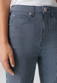Jeans en denim bleu et blanc rayés avec poches avant, quincaillerie visible et design ajusté, mettant en valeur des rayures verticales et une texture.