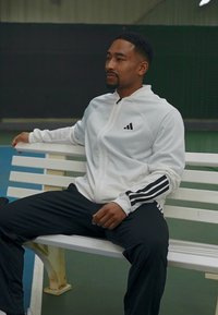 Witte zip-up hoodie met zwarte accenten, voorzien van een geborduurd logo, gecombineerd met zwarte trainingsbroek. Glad materiaal, ontspannen pasvorm, zittend op een bank.