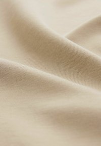 Beige bomuldsstof draperer blødt og viser en glat tekstur og fine ribbede mønstre med subtile variationer i lys og skygge.