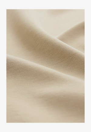 Un tessuto di cotone beige si drappeggia dolcemente, mostrando una consistenza liscia e un delicato motivo a coste, con sottili variazioni di luce e ombra.