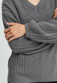 Grauer Pullover mit V-Ausschnitt, strukturiertem Strickmuster und gerippten Manschetten. Lockere Passform mit markantem Zopfmuster an Ärmeln und Körper.