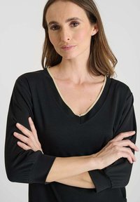 Donna con pelle chiara e capelli castani che indossa un top nero a V con rifiniture beige, braccia incrociate, espressione neutra, su uno sfondo neutro.