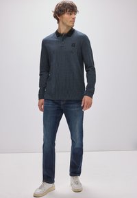 Dunkelblauses Langarmpolohemd mit strukturiertem Muster, schwarzem Kragen und Logodetail, kombiniert mit blauen Jeans und weißen Sneakers.