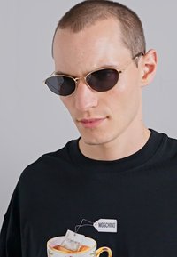 Sunglasses with black lenses and thin gold frames, worn with a black t-shirt featuring a tea cup graphic and a "Moschino" label.