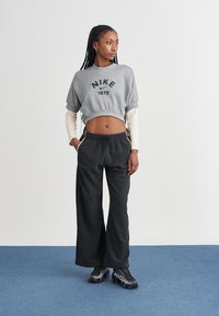 Šedá zkrácená mikina s potiskem "NIKE 1972", černé volně střižené kalhoty a černé tenisky s texturovanými detaily, na neutrálním pozadí.