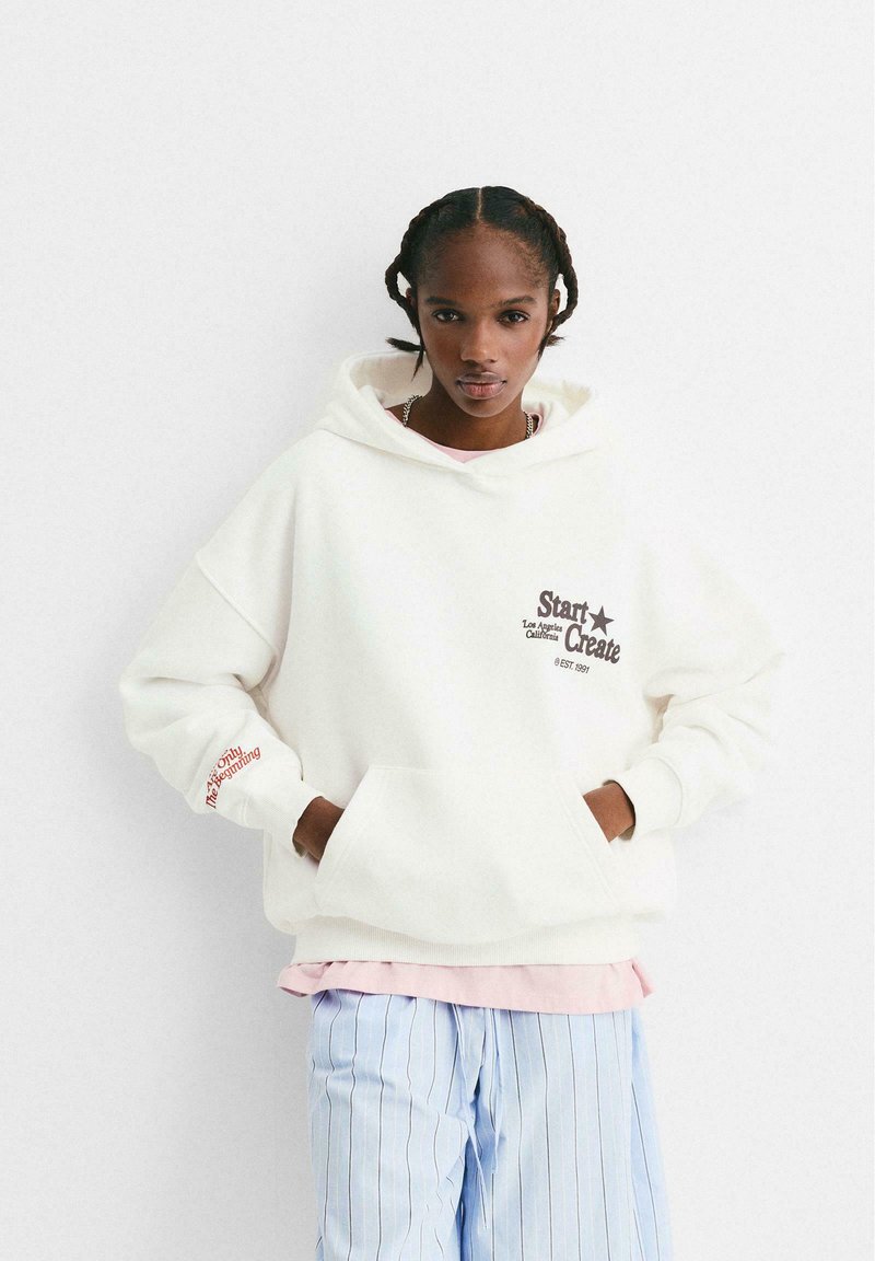 Ung person med flätat hår som bär en överdimensionerad vit hoodie med text och ljusblå randiga byxor, stående med händerna i fickorna.